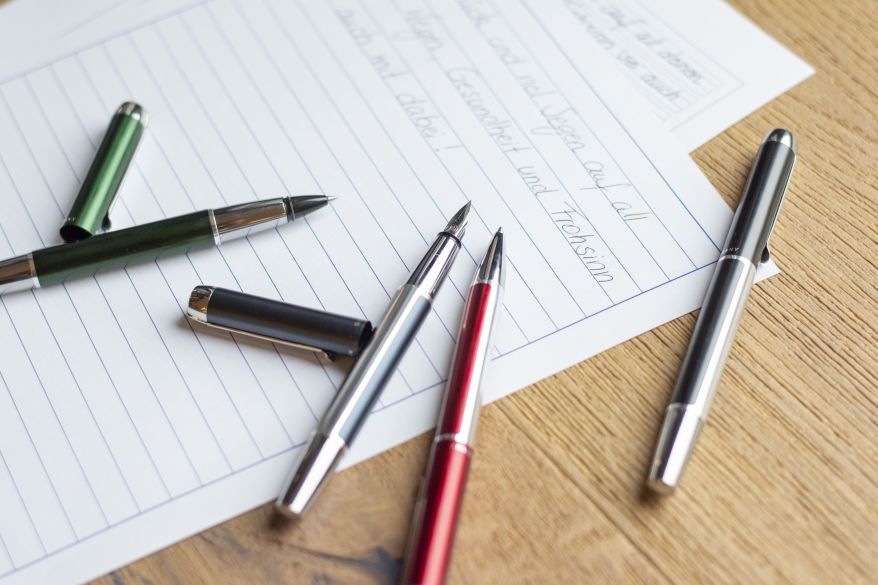 Füllhalter, Kugelschreiber und Tintenroller der Serie Pura von Pelikan liegen auf einem Schreibtisch auf zwei Blättern Papier.