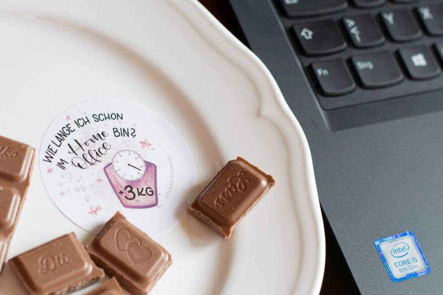 Nahaufnahme eines runden Aufklebers von Avery Zweckform auf dem steht: "Wie lange ich schon im Homeoffice bin? 3 Kilogramm." Man sieht einen Teller mit Schokoladenstücken und eine Tastatur.