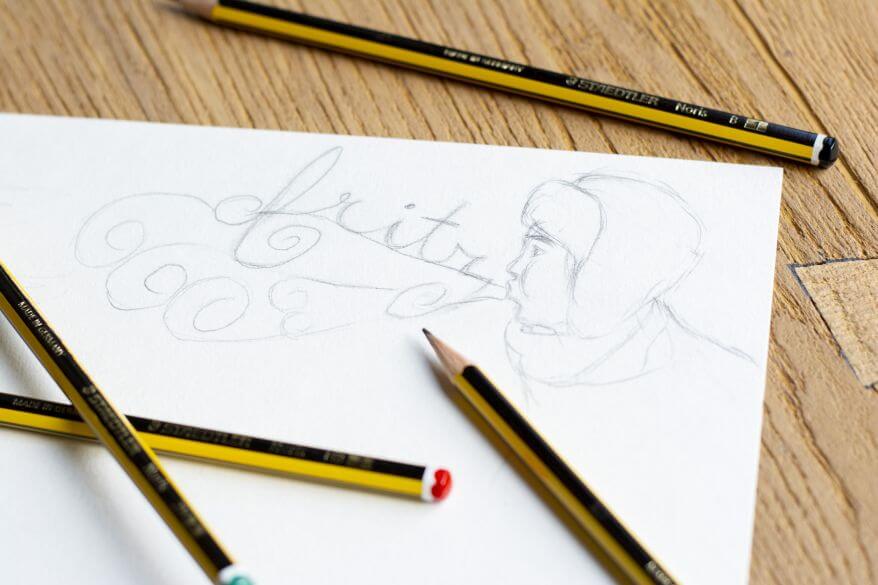 Nahaufnahme einer Bleistiftskizze: Ein Mann mit Chapka-Mütze pustet Wolken, in denen der Name "Fritz" steht. Um die Zeichnung herum liegen die Bleistifte Noris der Marke Staedtler.