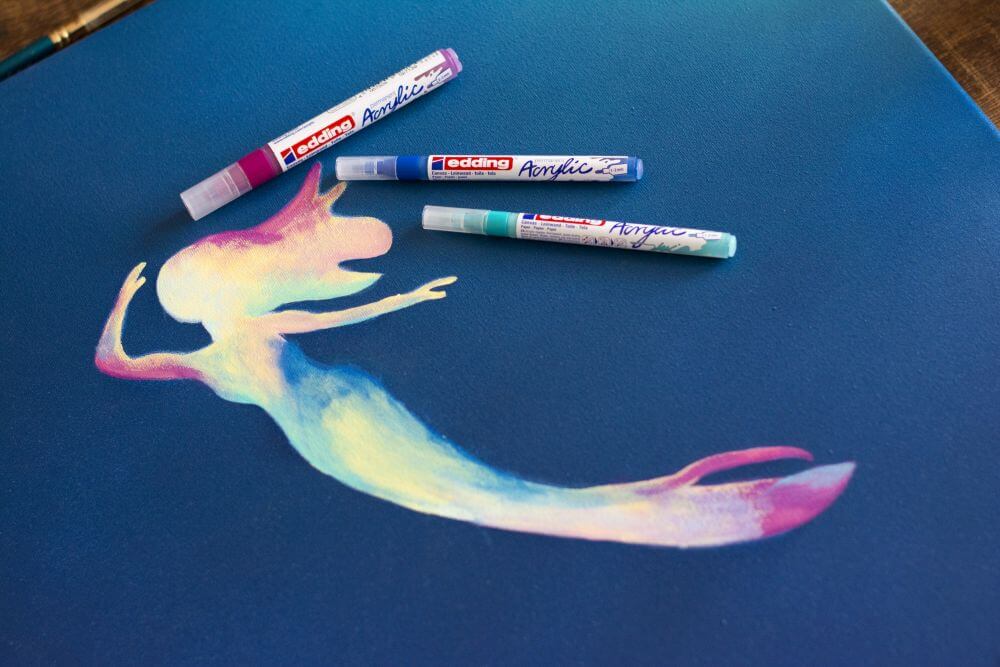Eine mit Acrylfarben gemalte Meerjungfrau ist auf einer Leinwand zu sehen. Um sie herum liegen Acrylmarker von Edding.