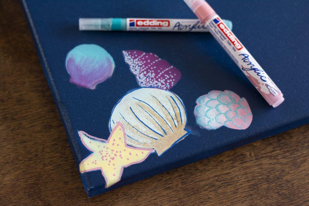 Nahaufnahme von Muscheln, Seestern und Seeschnecken, die mit Acrylmarkern von Edding auf eine Leinwand gemalt sind.