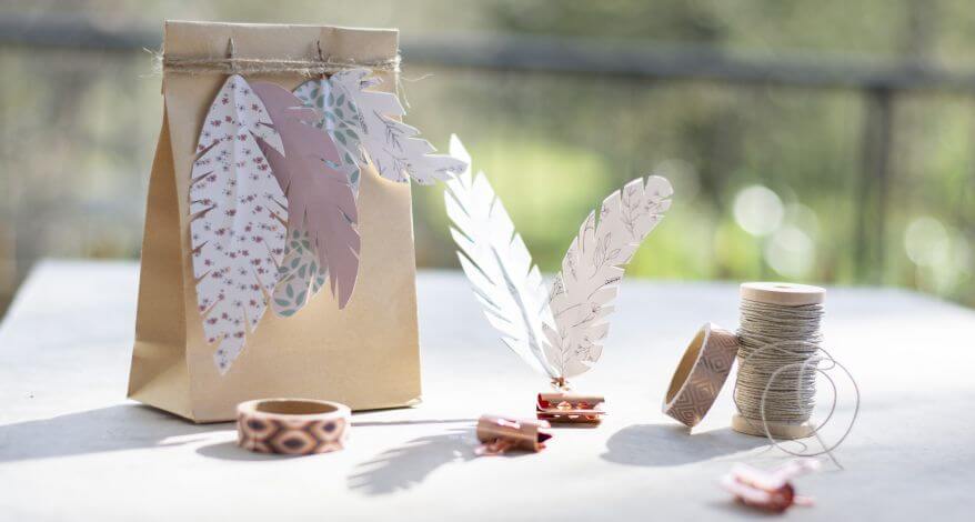 Auf einem Tisch steht eine Geschenktüte, an der mehrere Do it yoursefl-Papierfedern aufgehängt sind. Drumherum stehen Bastel- und Kreativmaterialien, die von der Sonne beschienen werden.