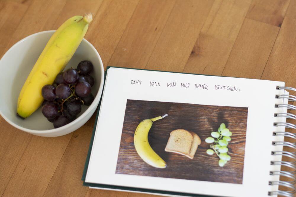 Ein Trostalbum für Kindergartenkinder: Ein Foto von einer Banane, einem Zwieback und Trauben ist mit dem Schriftzug "Damit kann man mich immer bestechen" in ein Fotoalbum Mini Mucho von Semikolon eingeklebt.