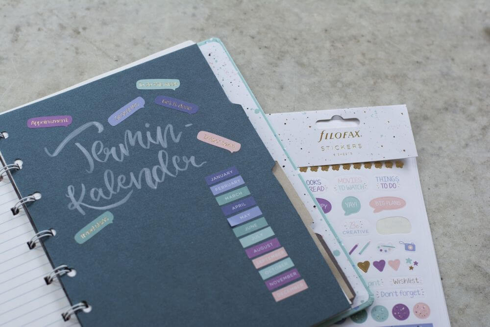 Ein Notizbuch von Filofax liegt aufgeklappt auf einem Schreibtisch, zu sehen ist ein selbstgemachter Divider mit dem Titel "Terminkalender". Auf das Papier sind verschiedene Aufkleber geklebt, ein Stickerbogen liegt im Hintergrund.