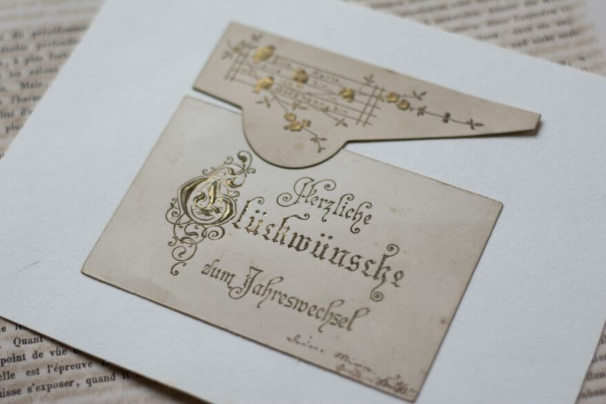 Ansicht einer historischen Postkarte mit Goldprägung. Auf der Grußkarte steht geschrieben "Herzlichen Glückwunsch zum Jahreswechsel".