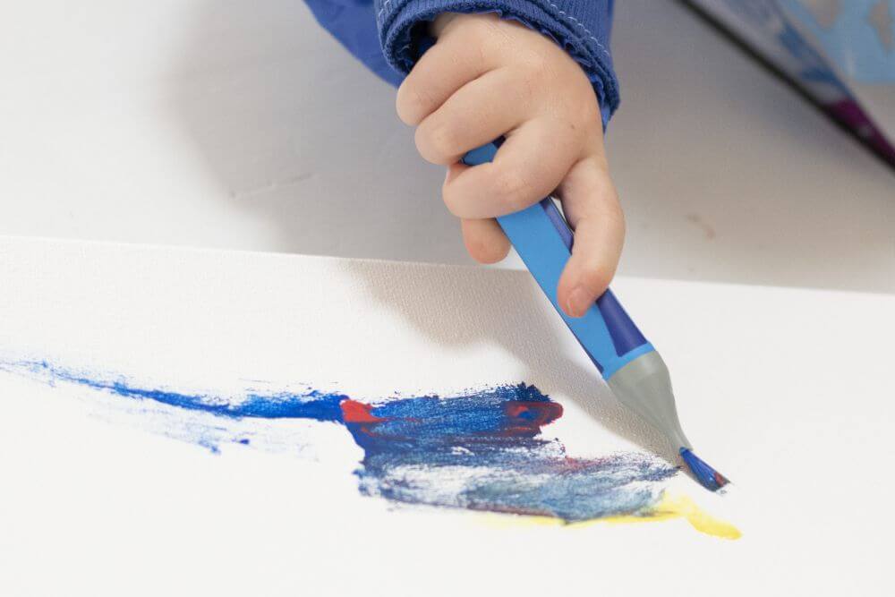Detailaufnahme einer Kinderhand, die mit Pinsel und Gouachefarben auf eine Leinwand malt.