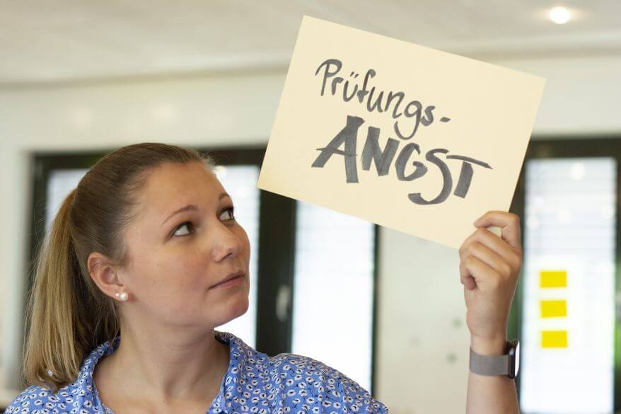 Eine junge Frau hält ein Schild hoch, auf dem das Wort "Prüfungsangst" steht. Sie betrachtet das Schild. Der Hintergrund ist hell und es sind unscharf Fenster zu erkennne. "