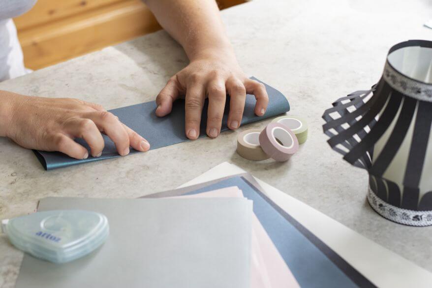 Hände falten einen DIN A4-Papierbogen Artoz in der Länge, um daraus eine Laterne aus Papier herzustellen.