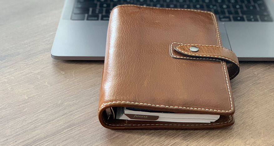Ein Kalender aus braunem Leder der Marke Filofax liegt auch einem Laptop.
