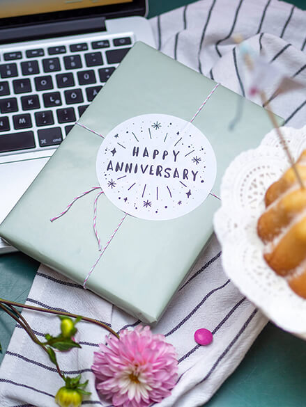 Ein Geschenk mit einem Sticker, auf dem "Happy Anniversary" steht, steht auf einem Laptop. Am Bildranf erkennt man einen Kuchen.
