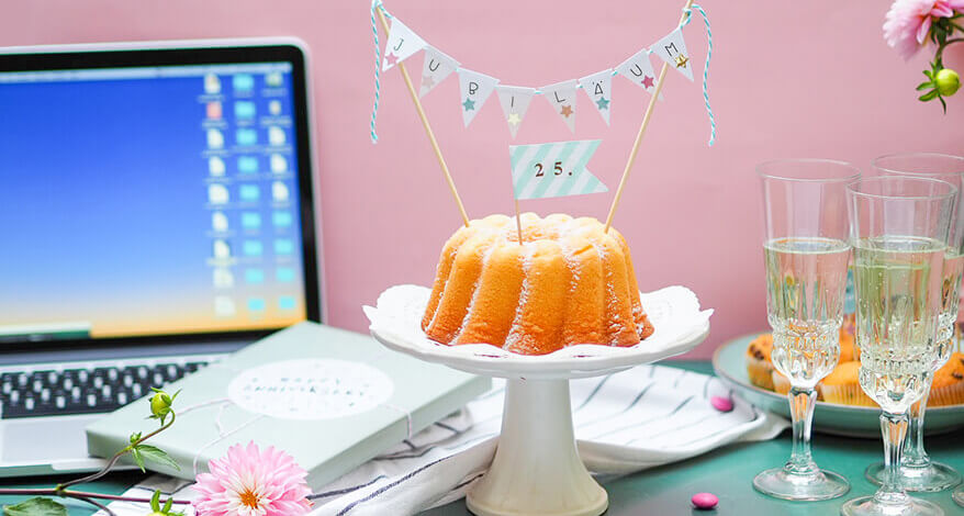 Ein Gugelhupf mit Jubiläums-Deko steht auf einer Tortenplatte, daneben stehen ein Laptop, Sektgläser und Blumen.