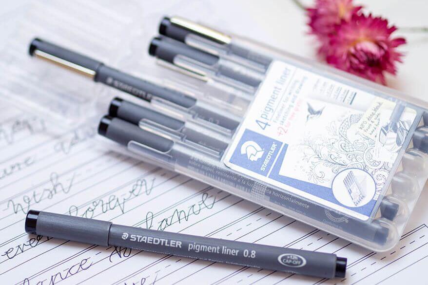 Auf einem Blatt mit Bounce Lettering Übungen liegt eine Box mit acht verschiedenen Pigment Linern der Firma Staedtler. Ein Stift ist herausgenommen und liegt vor dem Etui aus Plastik. Foto: Johanna Rundel