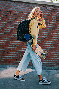 Ein Mädchen geht vor einer roten Mauer entlang und lächelt in die Kamera. In der Hand hält sie ein Longboard. Auf dem Rücken trägt sie einen Schulrucksack der Marke satch.