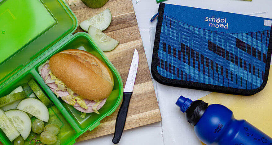 Auf einem Holzbrettchen liegt eine aufgeklappte Brotdose und eine Trinkflasche der Marke School-Mood. Die Snackbox ist mit einem belegten Brötchen, Gurkenscheiben und Trauben gefüllt. Neben dem Pausen-Frühstück sind ein Federmäppchen, Schulhefte und Buntstifte auf dem Tisch verteilt. Foto: Johanna Rundel