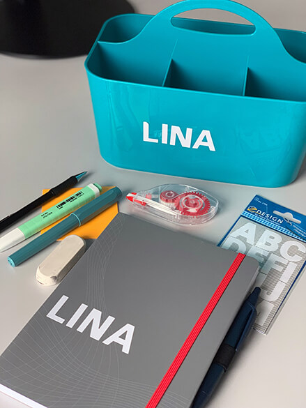 Auf einem Schreibtisch liegen verschiedene Büromaterialien: Ein Druckbleistift, ein Kaweco Füller, ein Textmarker und ein Korrekturmarker. Ein Notizbuch von der Marke Avery Zweckform wurde mit Buchstabenstickern beklebt. Darauf steht der Name Lina.