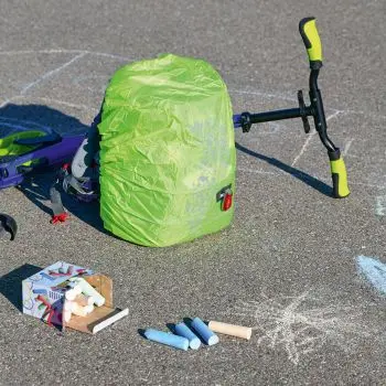 Auf einem asphaltierten Untergrund steht ein Schulranzen der Marke Step-by-Step mit fluoreszierender, gelber Regenhülle. Hinter dem Ranzen liegt ein umgekippter Roller, davor ein paar Straßenkreiden.