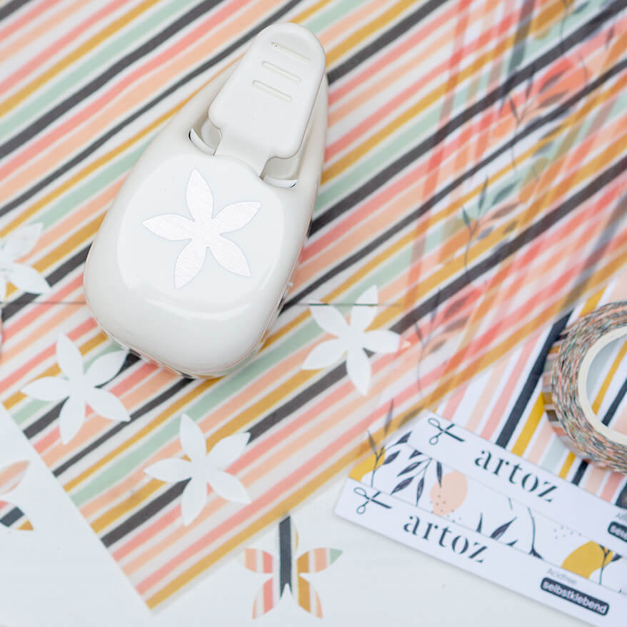 Ein Blumen-Stanzer der Marke Artoz steht auf ein paar Blättern Designpapier. Drumherum sind viele ausgestanzte Blümchen verstreut.