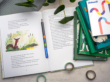 Auf einem grauen Untergrund liegt ein aufgeschlagenes Kinderbuch. In der Mitte liegt ein Lernbleistift der Marke Pelikan. Rechts neben dem Buch ist ein Stapel Bücher. Eine Pflanze ragt oben ins Bild. Ein paar Washitape-Rollen liegen lose verteilt herum. Foto: Johanna Rundel
