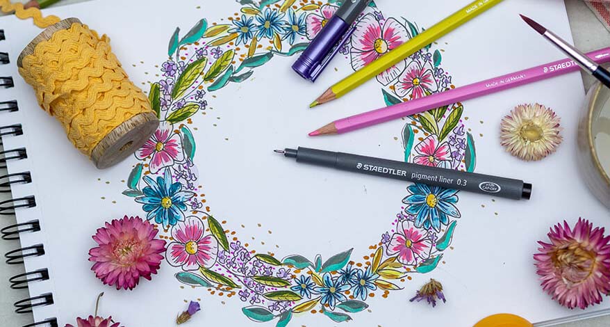 Auf einem Blatt Papier ist ein bunter Blumenkranz gemalt, darauf liegen Buntestifte, ein Fineliner und eine Rolle mit einer gelber Kordel. 