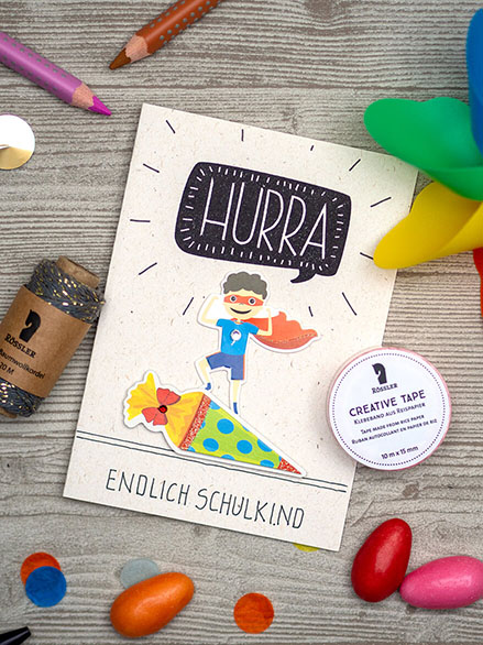 Auf einem hellgrauen Holzuntergrund liegt eine weiße Grußkarte mit einem gestempelten "Hurra". Außerdem befinden sich auf der Karte Aufkleber der Firma Rössler: ein Superhelden-Kind und eine Schultüte. Darunter der Spruch: "Endlich Schulkind". Drumherum liegen buntes Konfetti, eine Garnspule, Washitape und verschiedene Stifte verteilt.