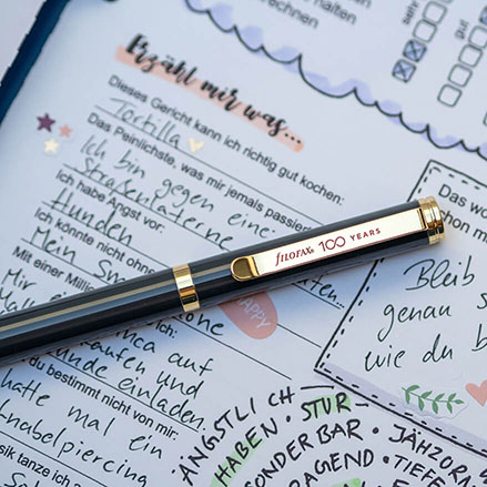 Auf einem hellgrauen Untergrund liegt ein aufgeschlagenes Clipbook von Filofax. Auf der Seite ist ein Teil eines ausgefüllten Freundebuches für Erwachsene zu sehen. Auf der Seite liegt ein dunkler Kugelschreiber mit goldener Klammer auf der "filofax 100 Years" steht. Foto: Johanna Rundel