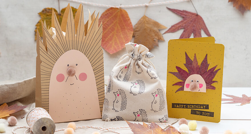 Auf einem hellen Untergrund, vor einer hellgrauen Betonwand, stehen eine zum Igel umgestaltete Papiertüte, ein zusammengebundenes Stoffsäckchen von Artoz, das mit kleinen Igel bestempelt wurde und eine gelbgrüne Klappkarte, auf der ein Igel aus Blättern aufgeklebt wurde. Drumherum und im Hintergrund sieht man viele getrocknete und gepresste Herbstblätter sowie kleine Pompons und eine Rolle Bäckergarn. Foto: Johanna Rundel 