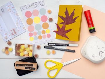 Auf einem hellen Untergrund liegen alle Bastelmaterialien, die man zu Herstellung einer Igel-Grußkarte benötigt: eine grüne Blanko Klappkarte, Stempel „Happy Birthday“´, ein schwarzes Stempelkissen von Artoz, getrocknete Herbstblätter, rosa Tonkarton, Klebestift, Marker in schwarz und weiß, Mini-Pompons, eine gelbe Schere, Klebestift und einen Stanzer. Foto: Johanna Rundel 