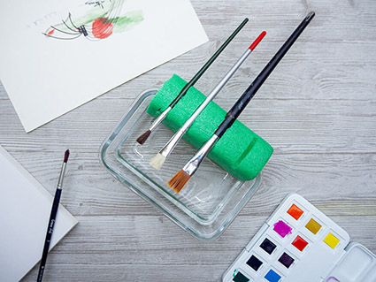 Auf einem hellgrauen Holzuntergrund steht ein Wasserbehälter an den seitlich ein Stück grüne Poolnudel geklemmt wurde, die drei Pinsel hält. Drumherum sind verschiedene Malutensilien verteilt.