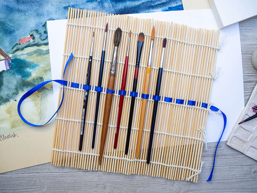 Auf einem hellgrauen Holzuntergrund liegt eine selbstgemachte Pinselrolle aus einer Bambusmatte und blauem Band. Untendrunter liegen einige fertige Bilder. Drumherum sind verschiedene Malutensilien verteilt.