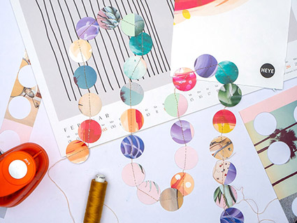 Auf einem hellen Untergrund liegt eine Girlande aus aneinander genähten Papierkreisen. Die Girlande besteht aus alten Kalenderblättern von Athesia. Außerdem sieht man Kalenderblätter mit Ausstanzungen, eine Stanze und eine Spule Garn. Foto: Johanna Rundel