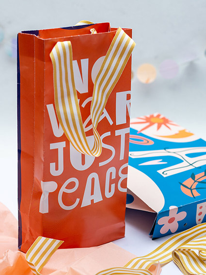 Zwei selbstgebastelte Papiertüten aus alten Kalenderblättern von Athesia. Die vordere ist orangefarbene mit gelb-weiß-gestreiftem Henkelband und steht aufrecht im Vordergrund. Darauf steht in weißer Schrift „No war just peace“. Dahinter liegt eine blaue Tüte mit buntem Muster. Foto: Johanna Rundel