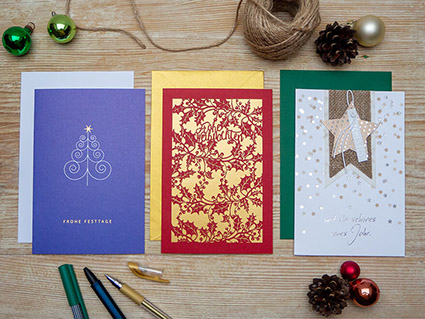 Auf einem Holztisch liegen verschiedene Weihnachtkarten von ABC Grußkarten. Links eine blaue, auf der in gold Frohe Weihnachten steht und ein abstrahierter Weihnachtsbaum abgebildet ist. In der Mitte liegt eine rot-goldene Karte mit weihnachtlichem Lasercut und rechts eine weiße Karte mit Golddetails, einem Jute Banner und einem ausgestanzten Stern. Drumherum sind weihnachtliche Deko, Juteband und Stifte drapiert. Foto: Johanna Rundel"