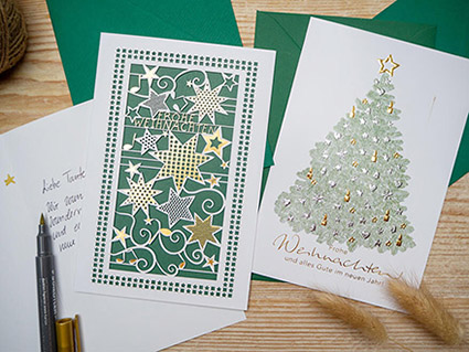 Auf einem Holztisch liegen verschiedene Weihnachtkarten von ABC-Grußkarten, sowie grüne Briefumschläge. Die Karten sind mit aufwändiger Goldprägung in unterschiedlichen Designs gestaltet. Ein geöffneter, goldener Brushpen liegt daneben. Foto: Johanna Rundel