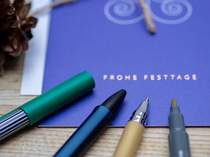 Auf einem Holztisch liegt eine blaue Weihnachtskarte von ABC Grußkarten, auf der in gold Frohe Weihnachten steht und ein abstrahierter Weihnachtsbaum abgebildet ist. Darunter liegen vier verschiedene Stifte. Ein grüner Fineliner, ein dunkelblauer Kugelschreiber, ein goldener Gelstift und ein Metallic-Brushpen in Gold. Foto: Johanna Rundel