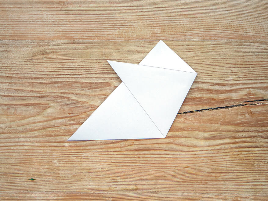 Ein zu einem Dreieck gefaltetes, quadratisches Blatt Papier der Marke Artoz. Eine Spitze des Dreiecks wurde schräg nach links oben gefaltet. Der Untergrund ist ein rustikales, helles Holzbrett. Foto: Johanna Rundel