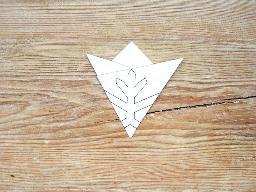 Auf die Spitze eines, zu einem Dreieck gefalteten Papier von Artoz, wurde der Umriss einer einfachen Schneeflocke gezeichnet. Der Untergrund ist ein rustikales, helles Holzbrett. Foto: Johanna Rundel