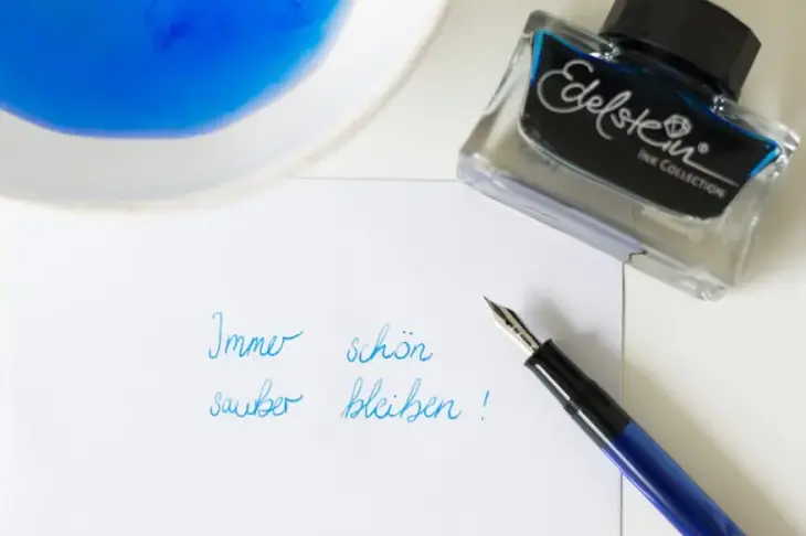Ein Blatt Papier, auf dem ein Füller liegt, ist beschrieben mit den Worten "Immer schön sauber bleiben". Im Hintergrund sind ein Schälchen mit Tintenwasser und ein Tintenglas Edelstein-Tinte zu sehen.