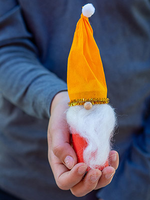 Ein Kind in einem dunkelgrauen Langarmshirt hält einen, aus einer leeren Klopapierrolle selbst gebastelten, Weihnachtswichtel mit orangefarbener Zipfelmütze aus einem Krepppapier von Heyda in die Kamera. Foto: Johanna Rundel