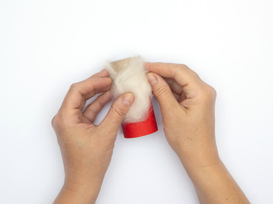 Jemand klebt weiße Filzwolle auf eine, mit einem Streifen roten Tonpapier von Heyda beklebte, leere Klopapierrolle. Der Untergrund ist weiß. Foto: Johanna Rundel