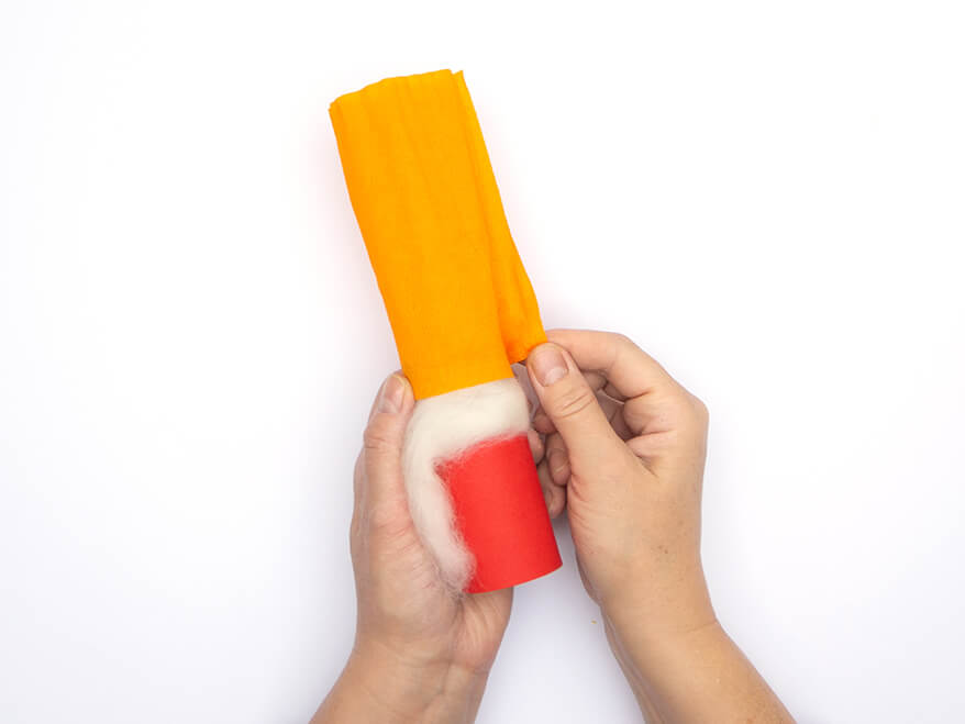 Jemand klebt einen Streifen orangefarbenes Krepppapier an den Rand einer leeren Klopapierrolle. Diese wurde zuvor mit weißer Filzwolle und einem Streifen roten Tonpapier von Heyda beklebt. Der Untergrund ist weiß. Foto: Johanna Rundel