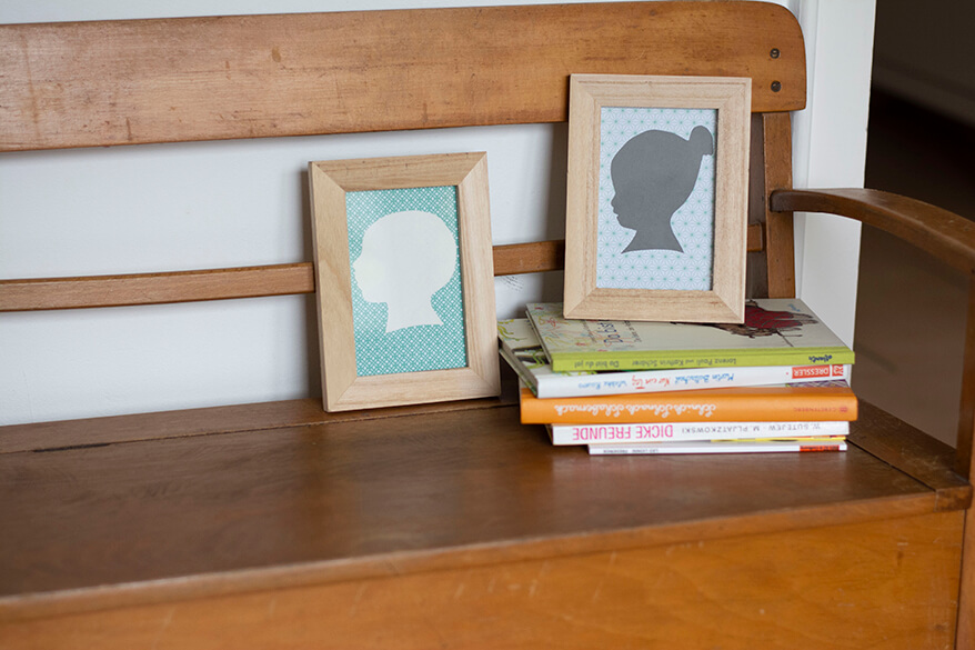 Auf einer hölzernen Kinderbank stehen die Do-it-yourself-Scherenschnitte zweier Kinderköpfe in zwei Bilderrahmen. Der Bilderrahmen mit dem Kopf eines Mädchens mit Dutt steht auf einem Stapel Kinderbücher, der Bilderrahmen mit dem Kopf eines Babys steht links davon direkt auf der Kinderbank.