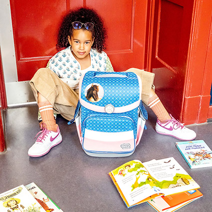 in Mädchen im Grundschulalter sitzt mit ihrem Schulranzen von School-Mood in den Armen auf dem Boden vor einer roten Tür. Das Mädchen hat seine Beine nach links und rechts ausgestreckt. Vor ihr liegen wahllos verschiedene Schulbücher. Das Mädchen hat dunkle Locken und eine Brille. Die Wand im Hintergrund ist dunkelblau.