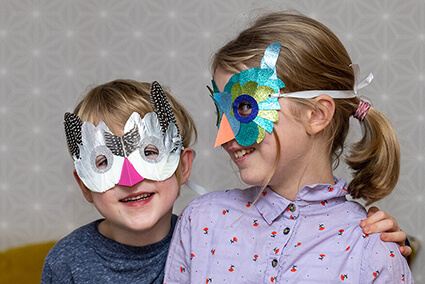Vor einem hellgrauen Hintergrund stehen zwei Kinder im Grundschulalter. Der Junge links trägt eine Vogelmaske mit echten Federn in Naturtönen mit pinkem Schnabel. Das Mädchen rechts hat eine Vogelmaske auf, die mit aquafarbenen Halbkreisen aus Glitzerpapier von Artoz beklebt wurde. Der Junge schaut in die Kamera, das Mädchen richtet den Blick seitlich auf den Jungen. Foto: Johanna Rundel