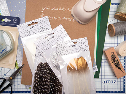 Auf einer Schneidematte von Artoz liegen verschiedene Bastelmaterialien, wie Papier in Grüntönen, Federn, eine Schere, helles Baumwollband, Kreisstanzer, Klebefilm und ein Stempel mit Federmotiv. Foto: Johanna Rundel