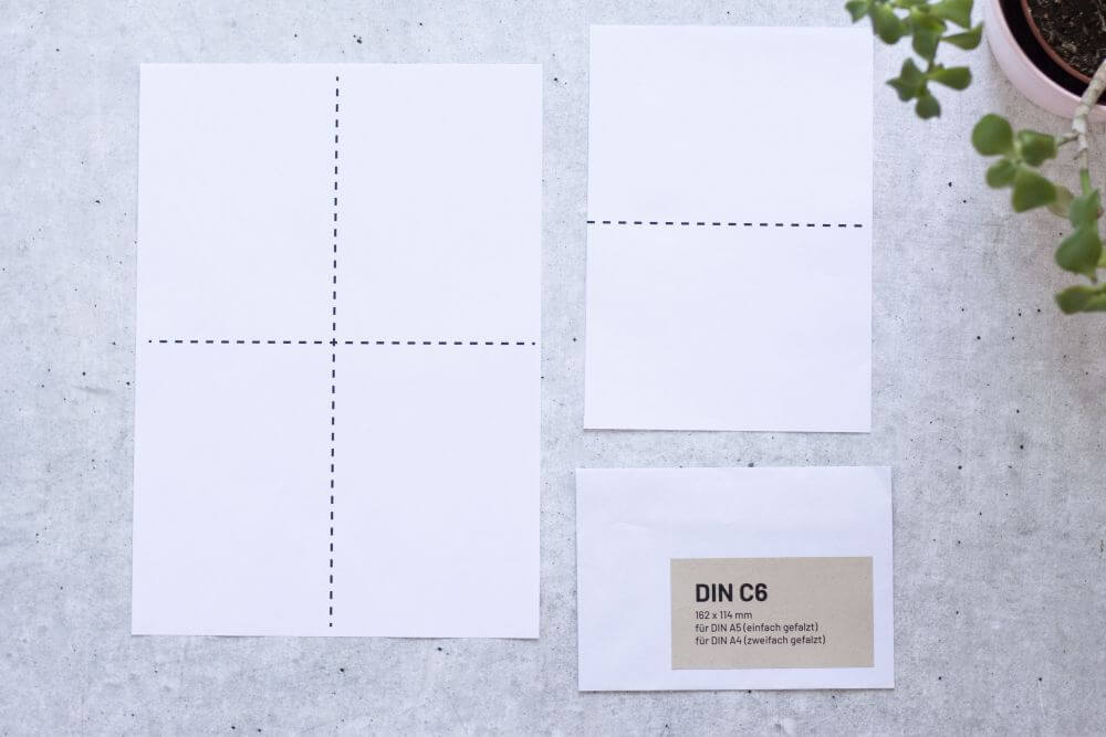 Auf grauem Grund liegt ein DIN A4-Papier mit zwei gestrichelten Markierungen, die das Papier in Viertel teilt und ein DIN A5-Papier mit einer gestrichelten Markierung in der Mitte, dazu ein passender Briefumschlag C6. Auf dem Briefumschlag ist ein Aufkleber aufgebracht mit folgenden Infos: DIN C6, 162x114 Millimeter, für DIN A5 (einfach gefalzt), für DIN A4 (zweifach gefalzt). Rechts oben ragt der Teil einer Pflanze ins Bild.