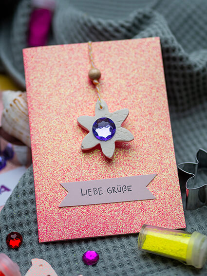 Auf einem salbeigrünen Küchenhandtuch liegt eine Grußkarte aus Glitterkarton von Heyda. Auf der Karte ist ein Anhänger aus Modelliermasse in Blumenform mit einem blauen Glitzerstein von Knorr Prandell zu sehen. Darunter liegt ein fliederfarbener Zettel, auf dem "Liebe Grüße" gestempelt worden ist. Vor der Karte liegt ein Glitterdöschen mit neongelbem Glitter und ein paar bunte Glitzersteine. Foto: Johanna Rundel 