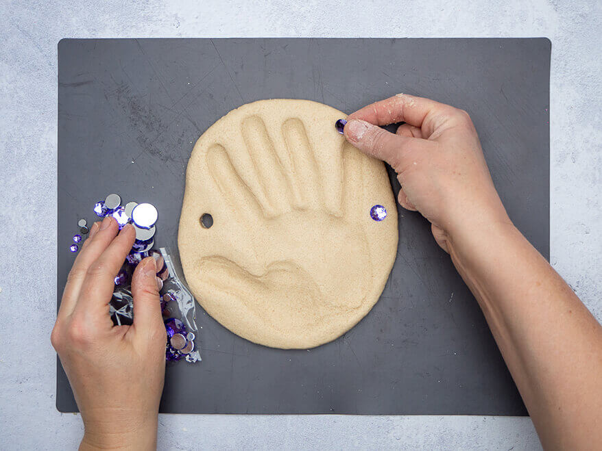 Jemand drückt Glitzersteine von Knorr Prandell in einen Kreis aus Salzteig, der auf einer dunkelgrauen Backmatte liegt. Der Untergrund ist Betongrau. Der Untergrund ist Betongrau. Foto: Johanna Rundel 