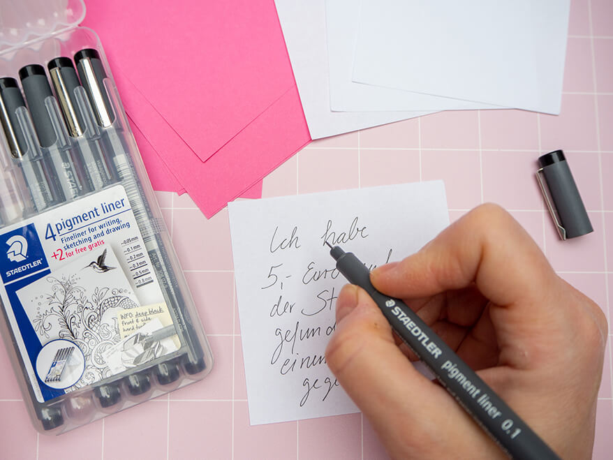 Jemand schreibt mit einem schwarzen Pigmentliner von Staedtler einen Glücksmoment auf einen weißen Haftnotizzettel. Auf dem Bild sind weitere Zettel in Pink und Weiß, sowie eine geöffnete Packung Pigmentliner in unterschiedliche Stärken zu sehen. Der Untergrund ist rosafarben mit weißem Karomuster. Foto: Johanna Rundel