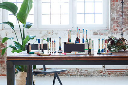 Viele verschiedene Stifte, Marker und Nachfüllkartuschen der EcoLine von edding stehen und liegen auf einem dunklen Holztisch. Im Hintergrund sind ein schwarzer Stuhl und weiße Fenster zu sehen. Auf und neben dem Tisch stehen zwei Pflanzen.