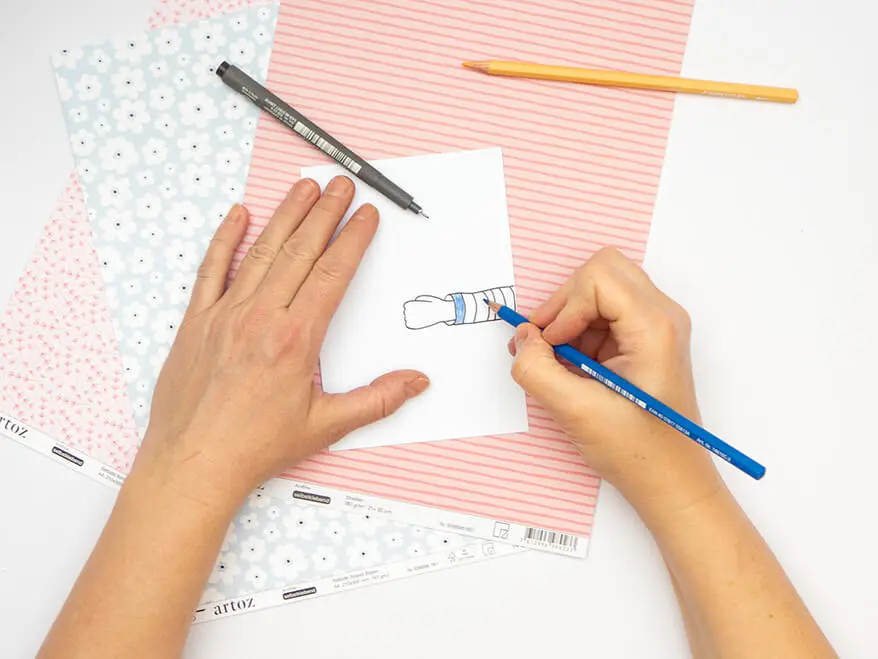 Jemand malt mit einem blauen Buntstift einen gezeichneten Arm aus, der seitlich in eine weiße Klappkarte von Artoz hineinragt. Unter der Klappkarte liegen weitere Designpapiere von Artoz. Der Untergrund ist weiß. Foto: Johanna Rundel